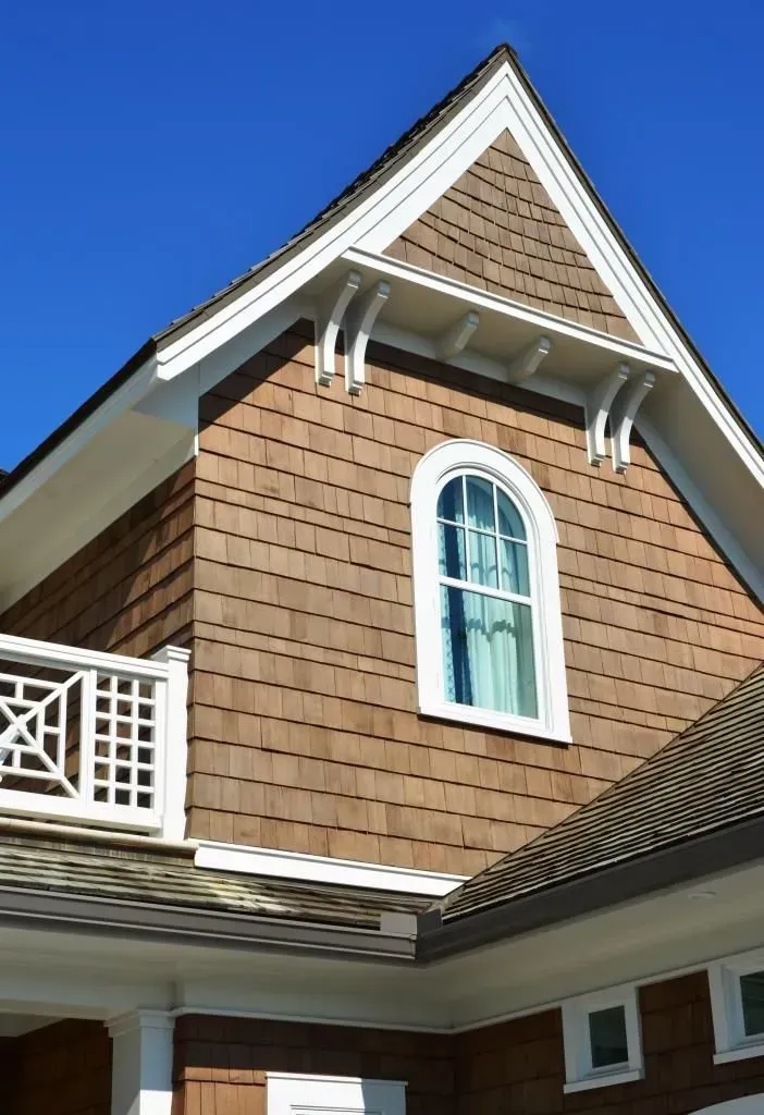 Brown shingled house exterior with white trim, arched window, and bracket details against a blue sky.