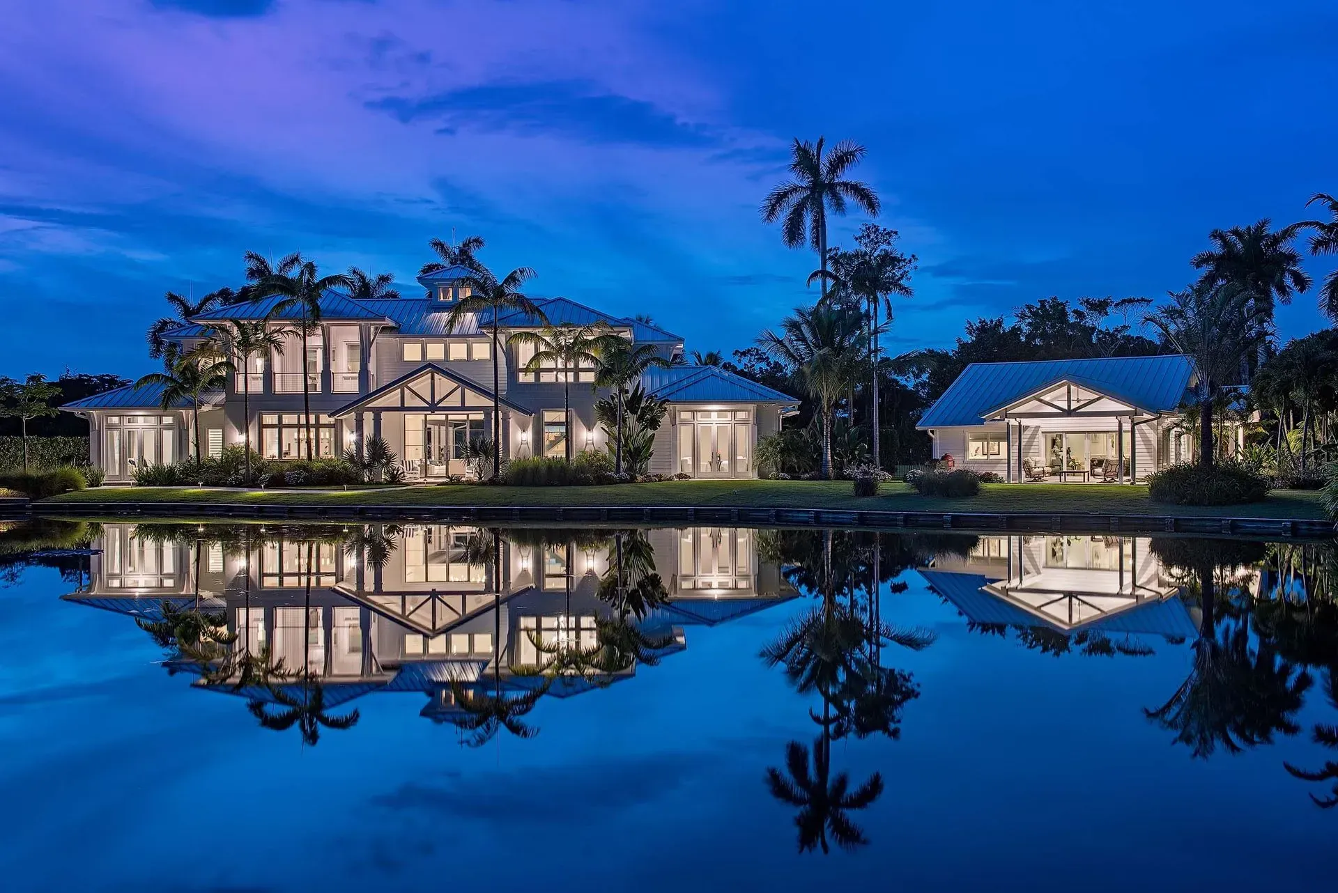 Large waterfront home with reflections in water at dusk; palm trees.