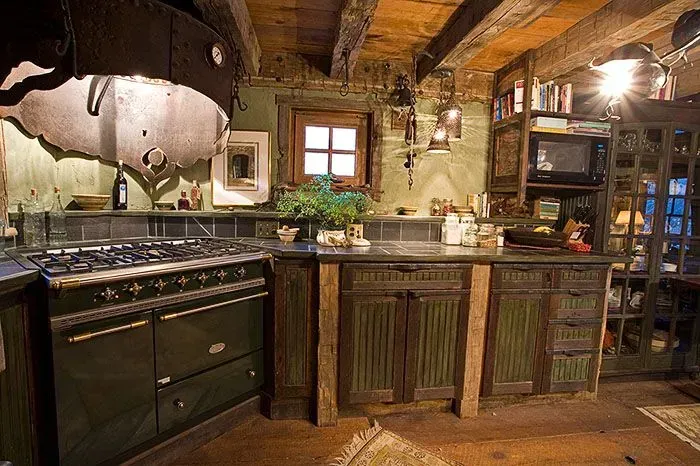 Rustic green kitchen with stove, cabinets, and small window. Wooden beams and shelves.