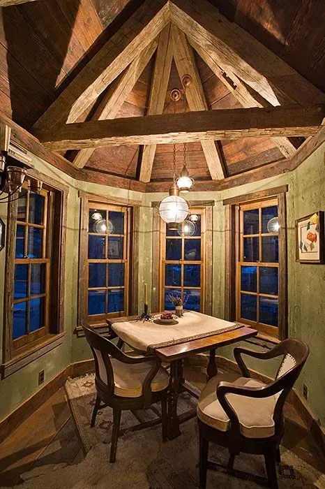 Rustic dining nook with wood beams, three windows, table, chairs, and hanging light fixture.