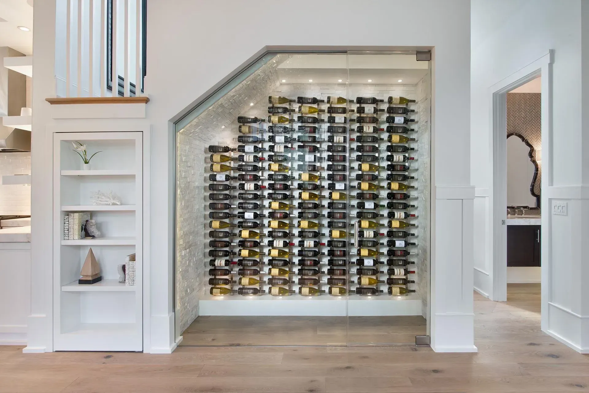 Built-in wine rack under stairs; clear glass walls; many wine bottles; white trim; light wood floor.