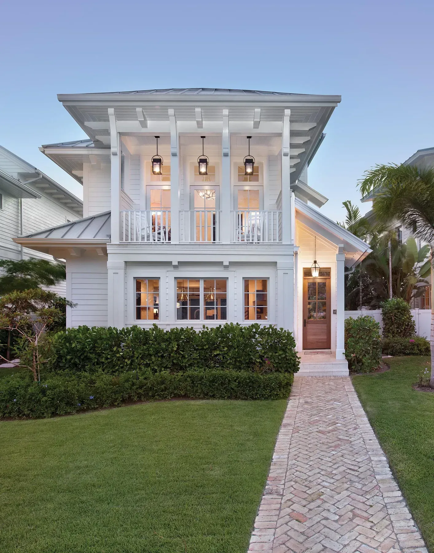 White two-story house with a brick walkway and green lawn. Balcony with hanging lights.