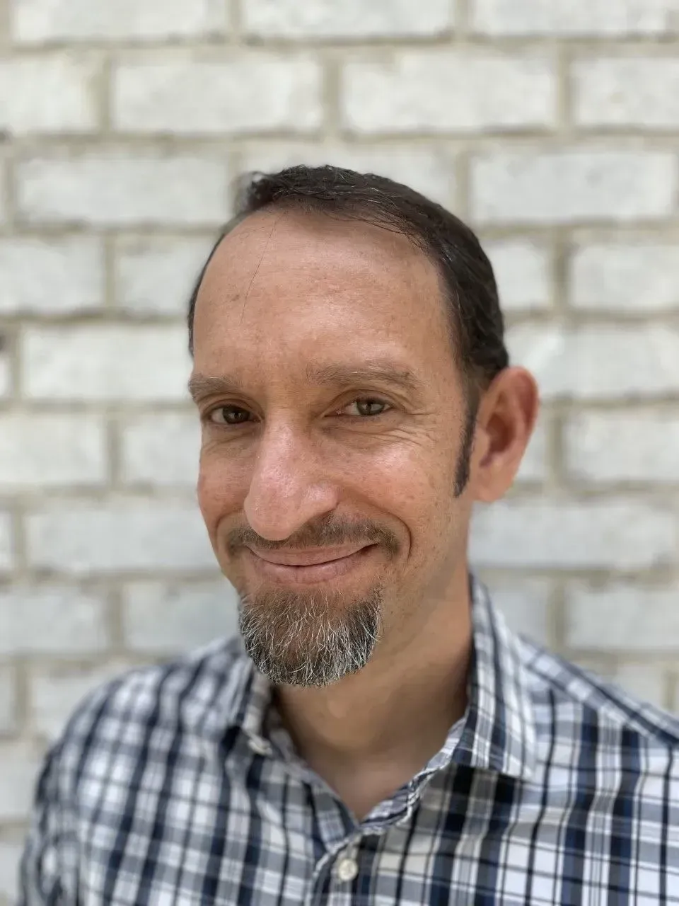 Man with a goatee smiling, wearing a plaid shirt, standing in front of a white brick wall.