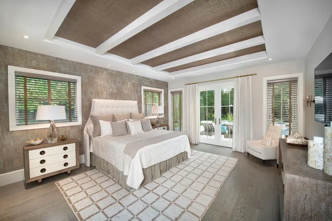 Bedroom with neutral tones; textured walls, coffered ceiling, bed, windows, rug.