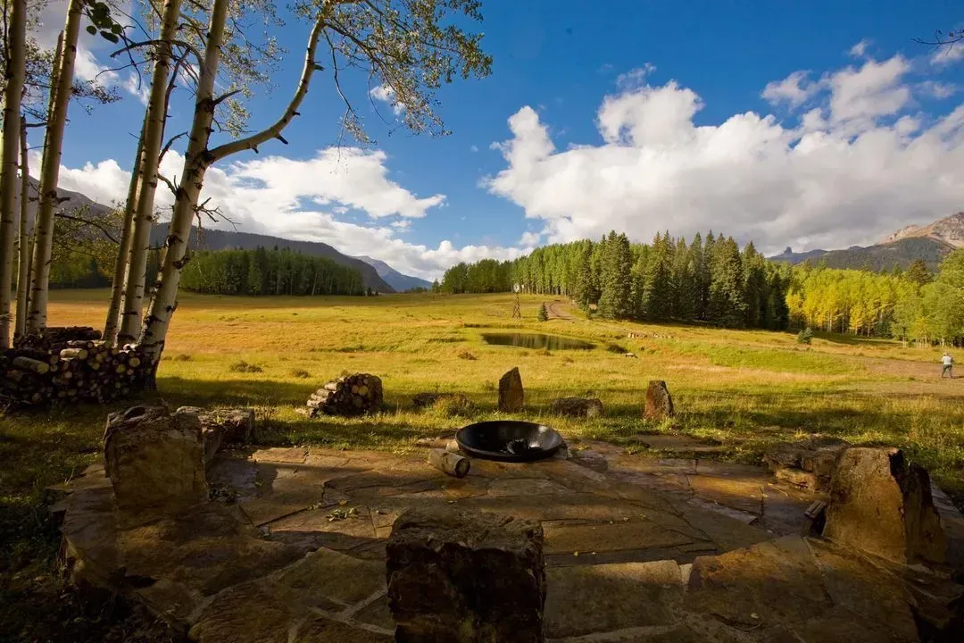 Campground with fire pit, grassy meadow, and trees under a blue sky with clouds.