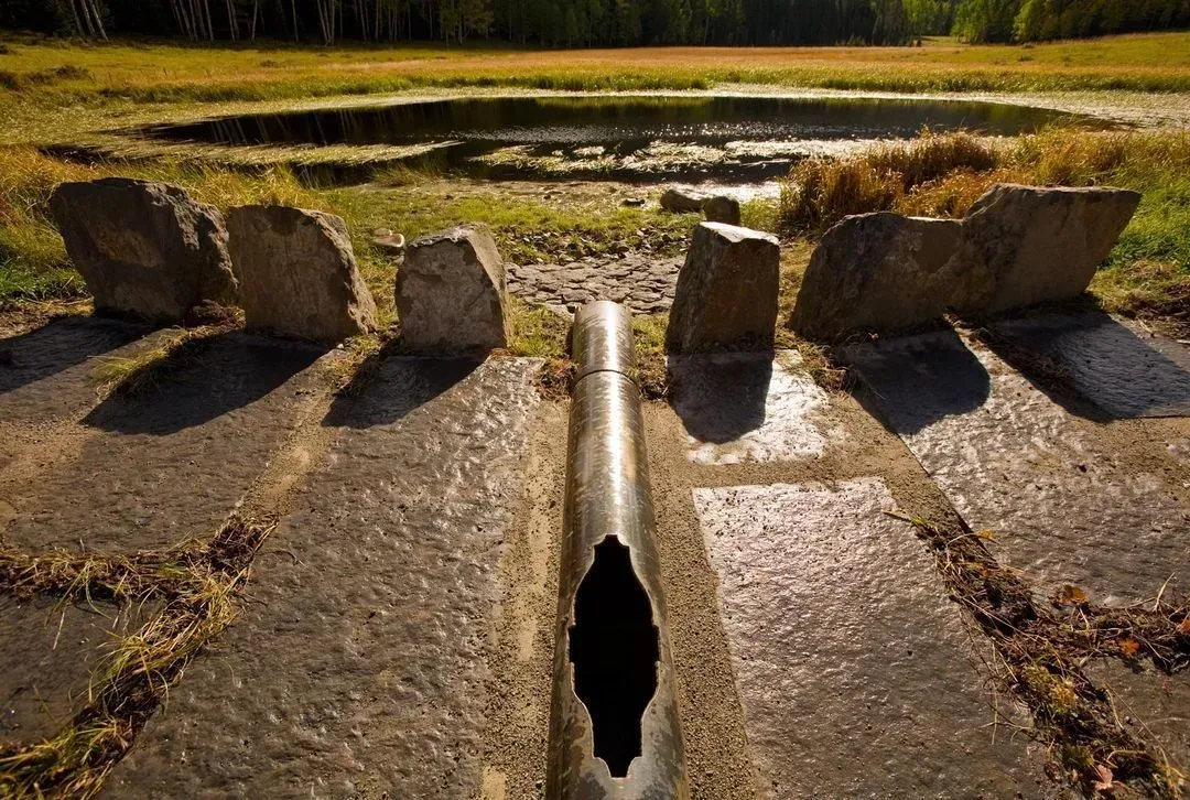 Stone latrines in a grassy field with a small pond in the background. Sunny day.
