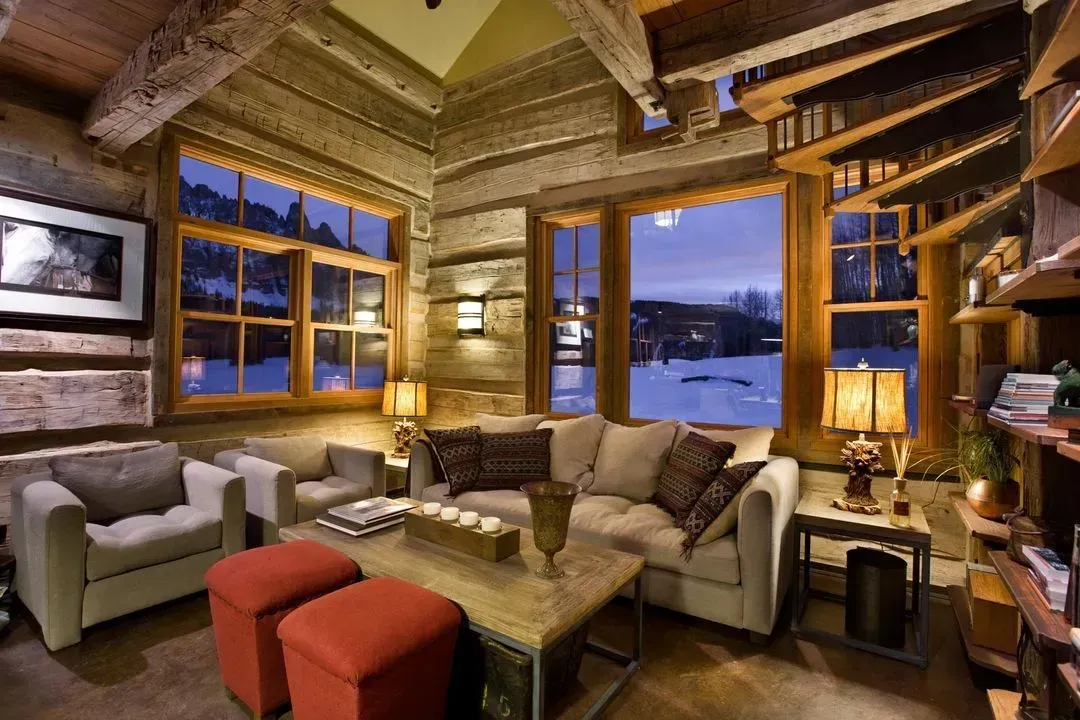 Cozy living room with log walls, large windows showing a snowy landscape, and warm lighting.