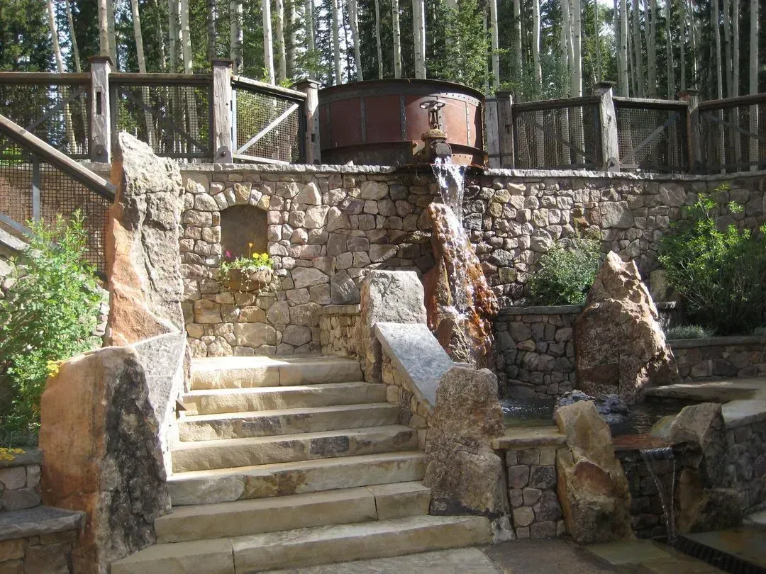 Stone staircase leading to a tiered water feature with a rusted metal container; wooden fence in the background.