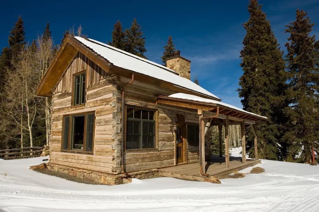 Log cabin in snow with porch, trees, and blue sky.