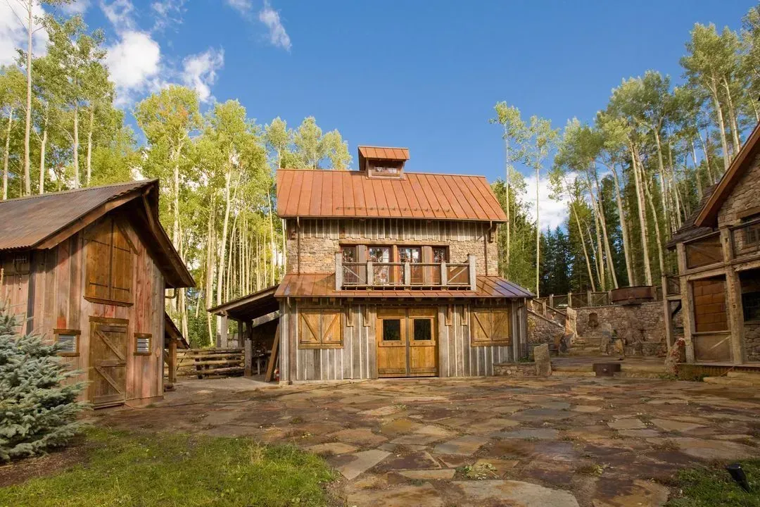 Rustic wooden buildings with rusty metal roofs set in a clearing, surrounded by trees under a blue sky.