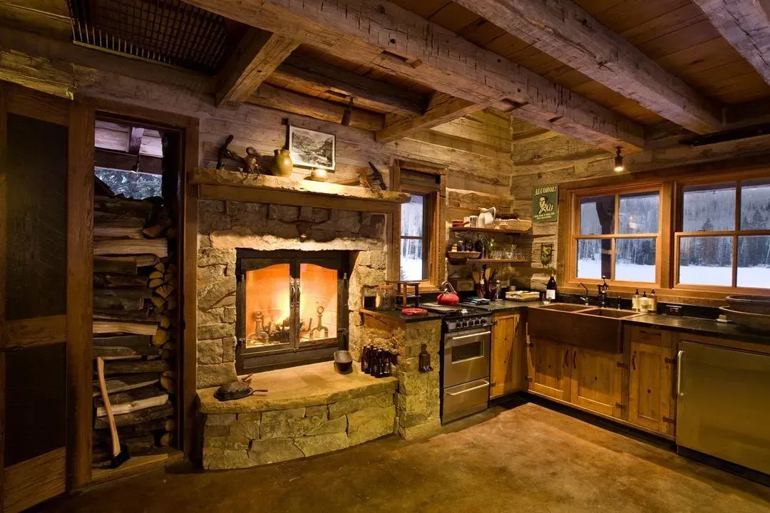 Cozy cabin kitchen with lit fireplace, wooden beams, stone hearth, and snowy windows.