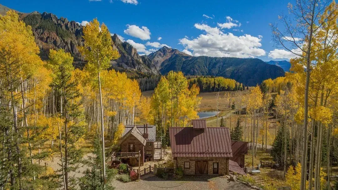 Cabin in autumn setting, surrounded by golden trees, lake, and mountains.
