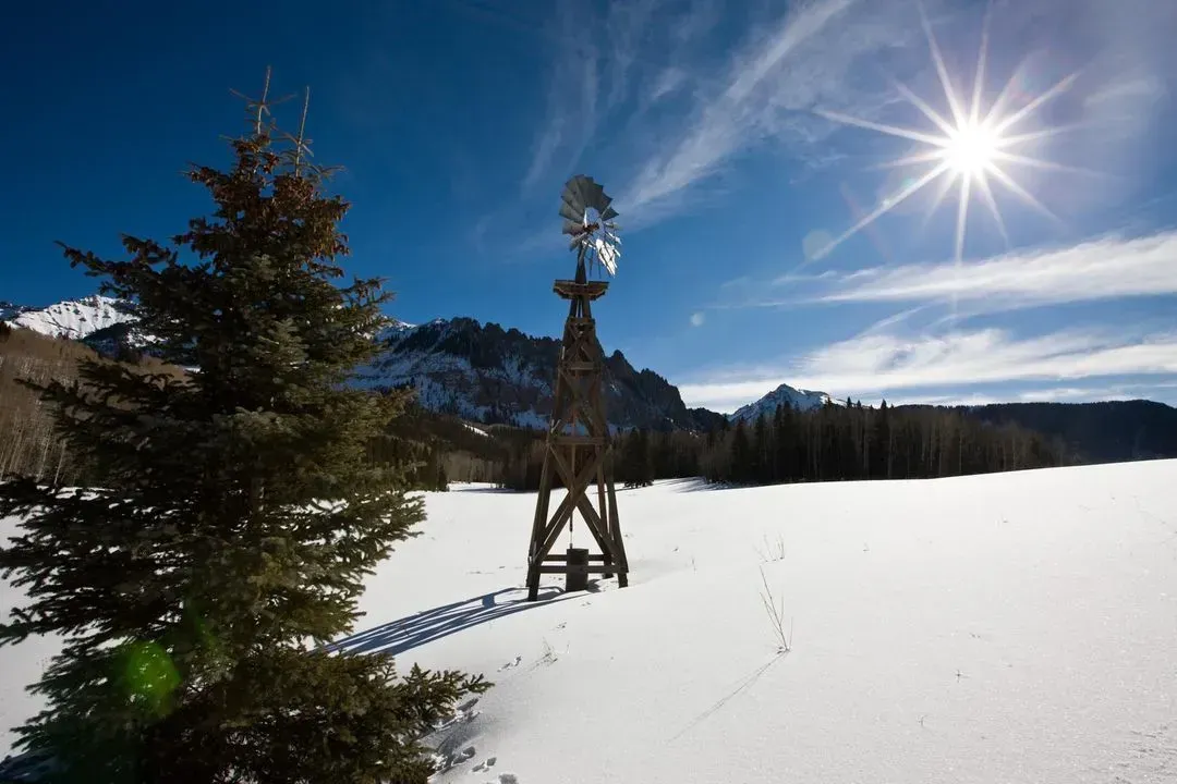 Snowy field with a windmill, pine tree, mountains, and bright sun in a blue sky.