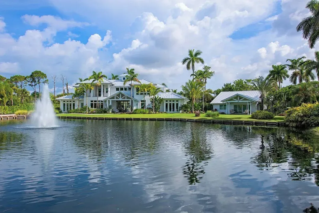 White mansion overlooking a lake with a fountain, surrounded by palm trees under a blue sky with clouds.