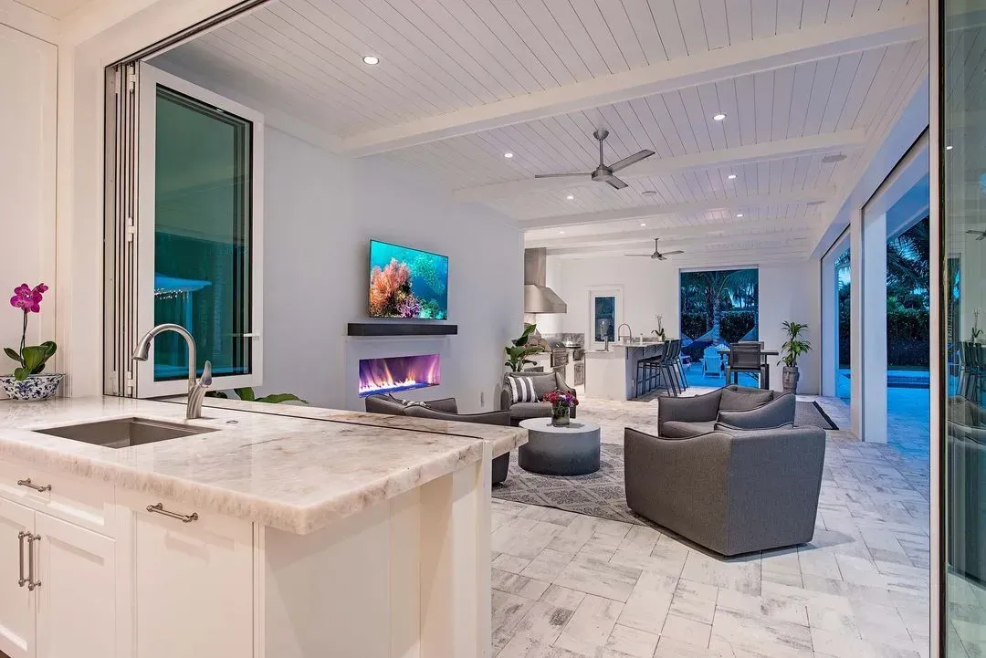 Open-concept kitchen and living area with folding glass doors. White cabinets, marble countertops, and a fireplace.