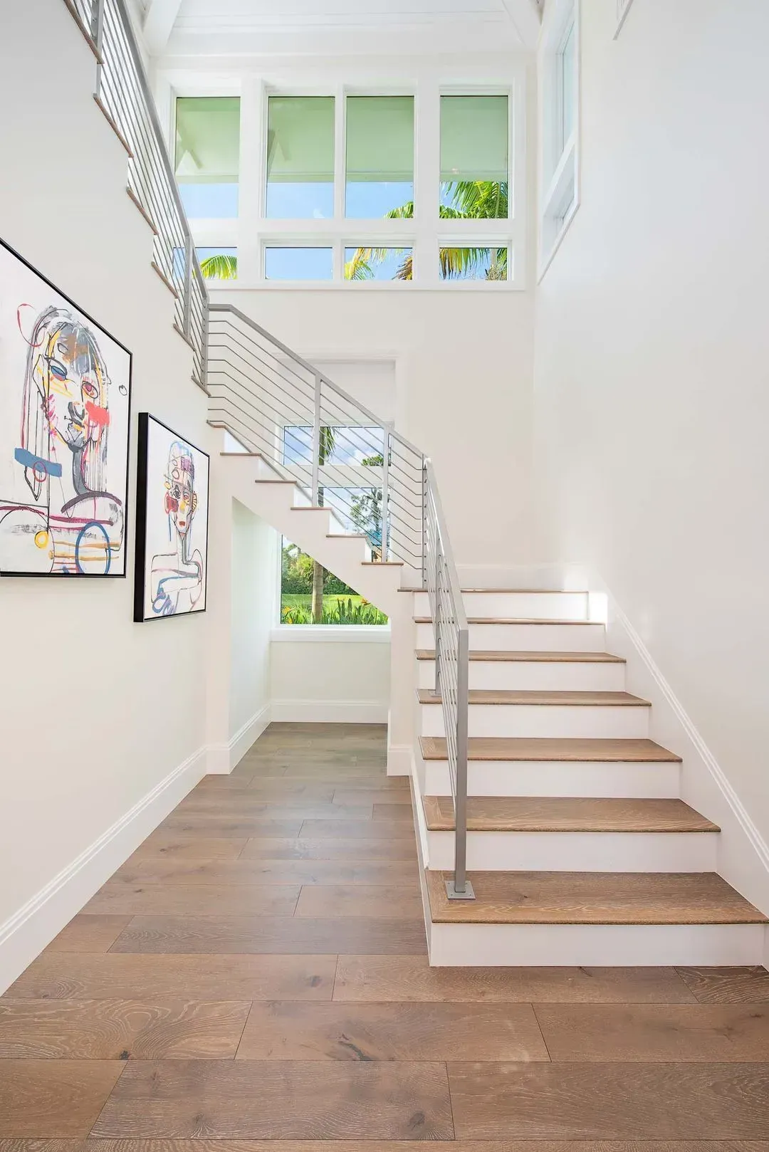 White interior with wooden staircase, metal railing, art on the wall, and windows above.