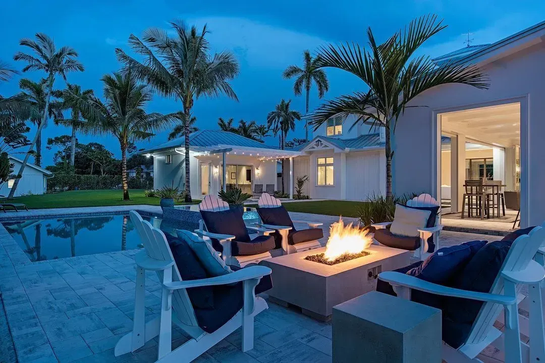 Nighttime outdoor patio with fire pit, seating, pool, and white house with palm trees.