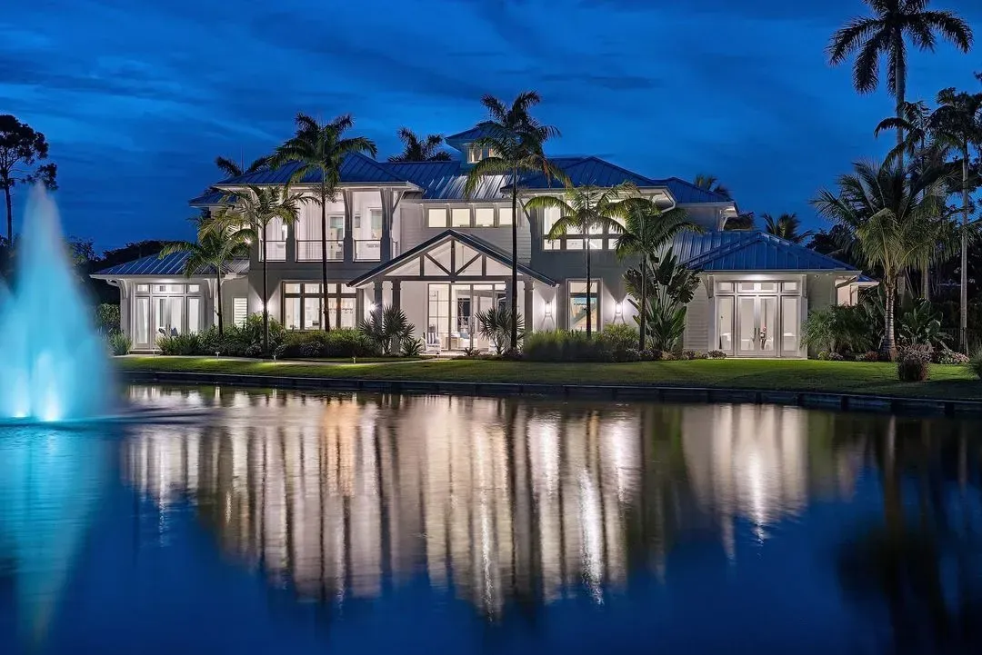 Luxury home at night, reflecting in a dark pond. Palm trees, fountain, and lights enhance the scene.