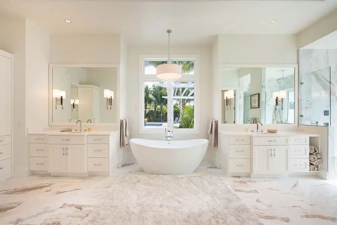 Bright, modern bathroom with a freestanding tub, dual vanities, large window, and marble flooring.