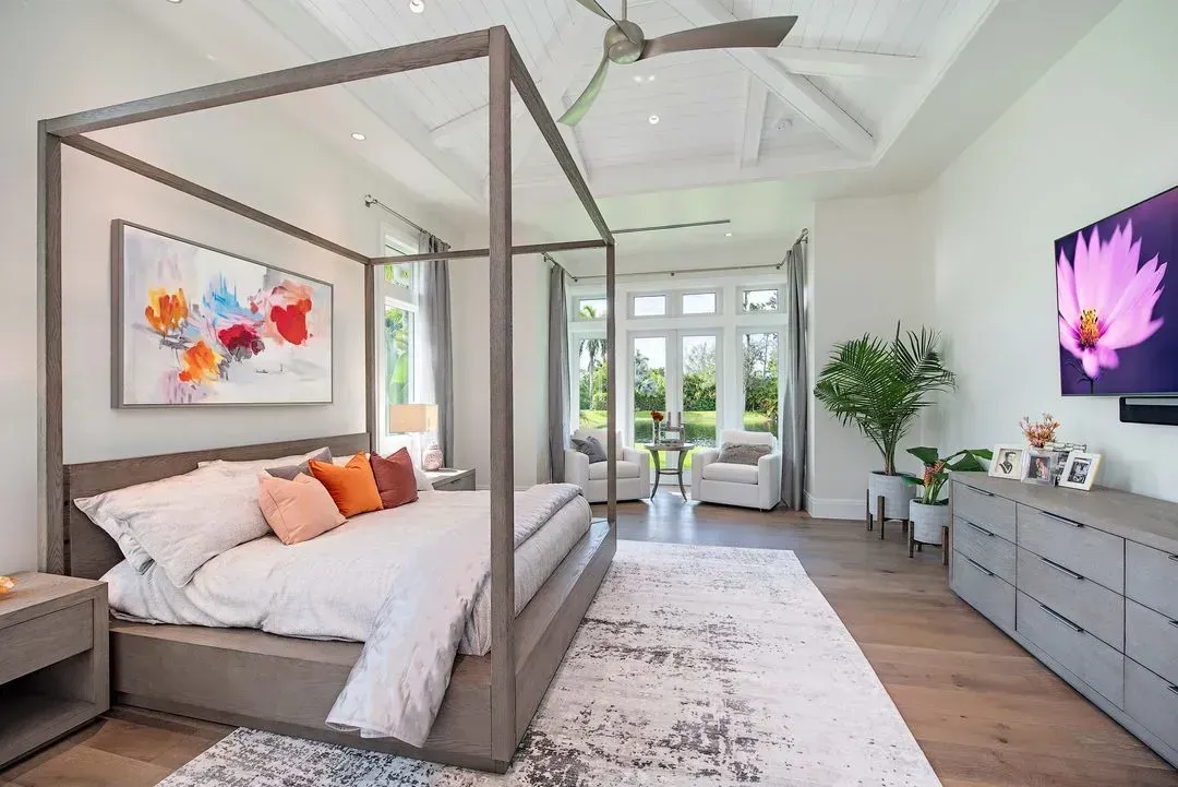 Spacious modern bedroom with canopy bed, artwork, TV, and outdoor access. Neutral colors, wooden floors.