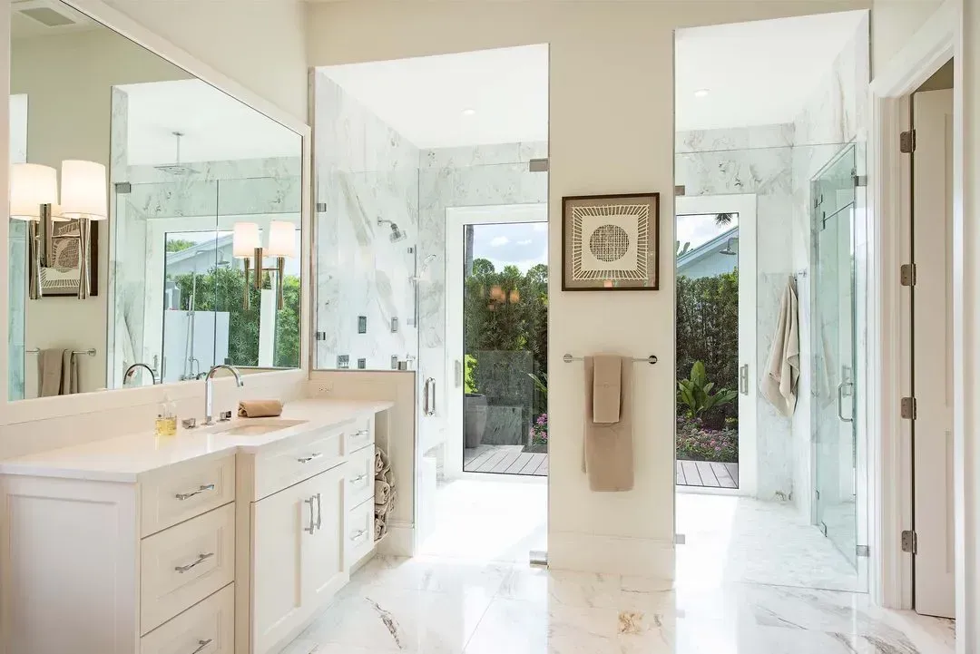 Luxurious white marble bathroom with a large vanity, glass shower, and open door to a backyard.