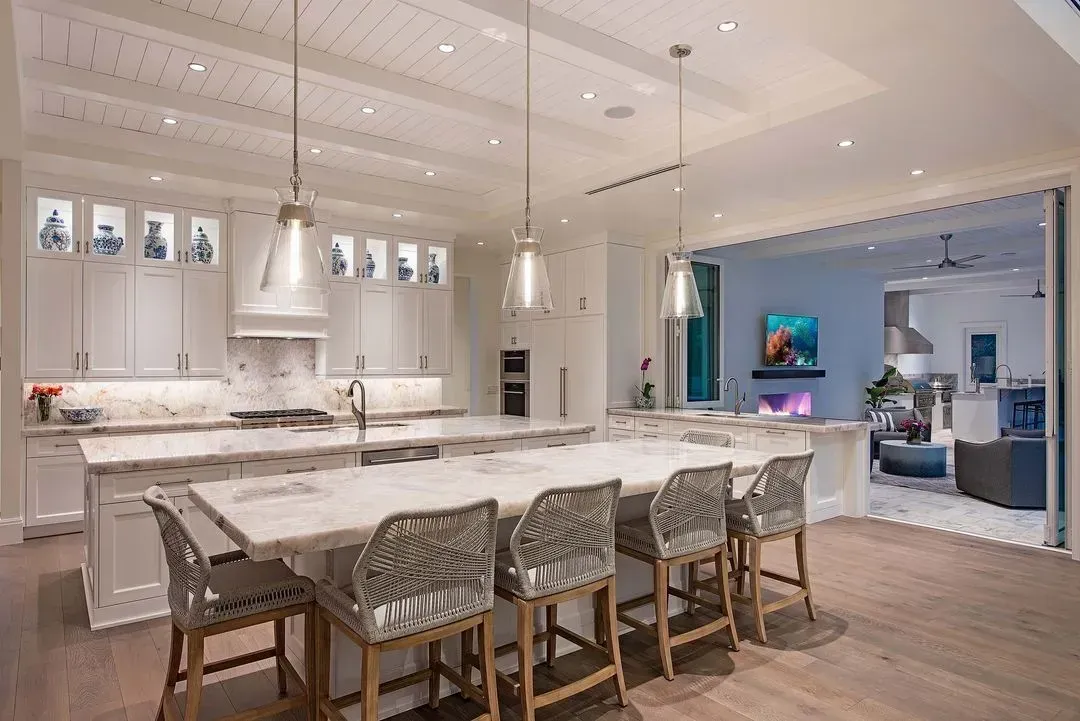 Elegant white kitchen with marble countertops, island seating, and open doorway to living area.