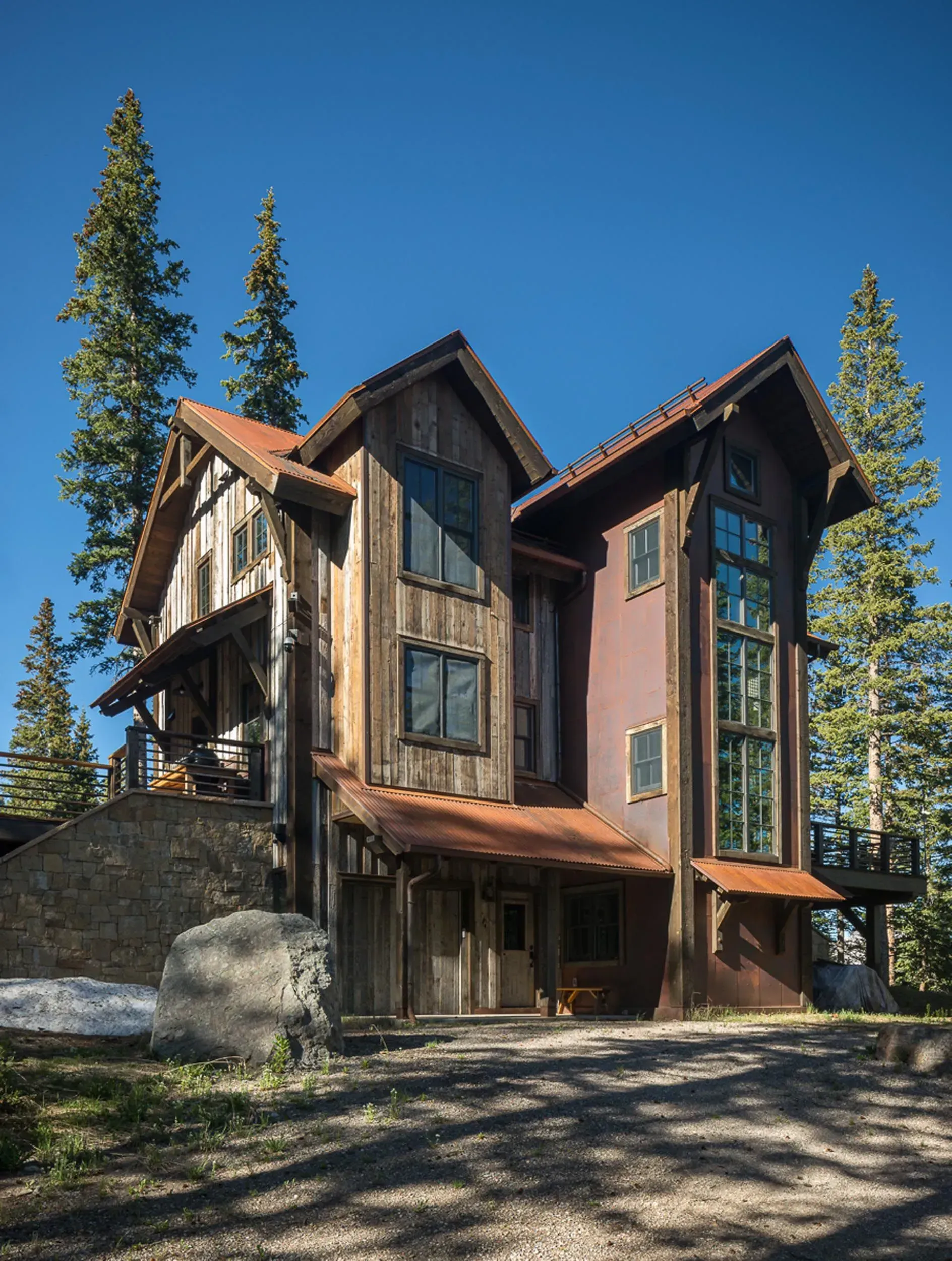 Rustic three-story mountain home with wood siding and a copper roof, set against a backdrop of trees and a blue sky.