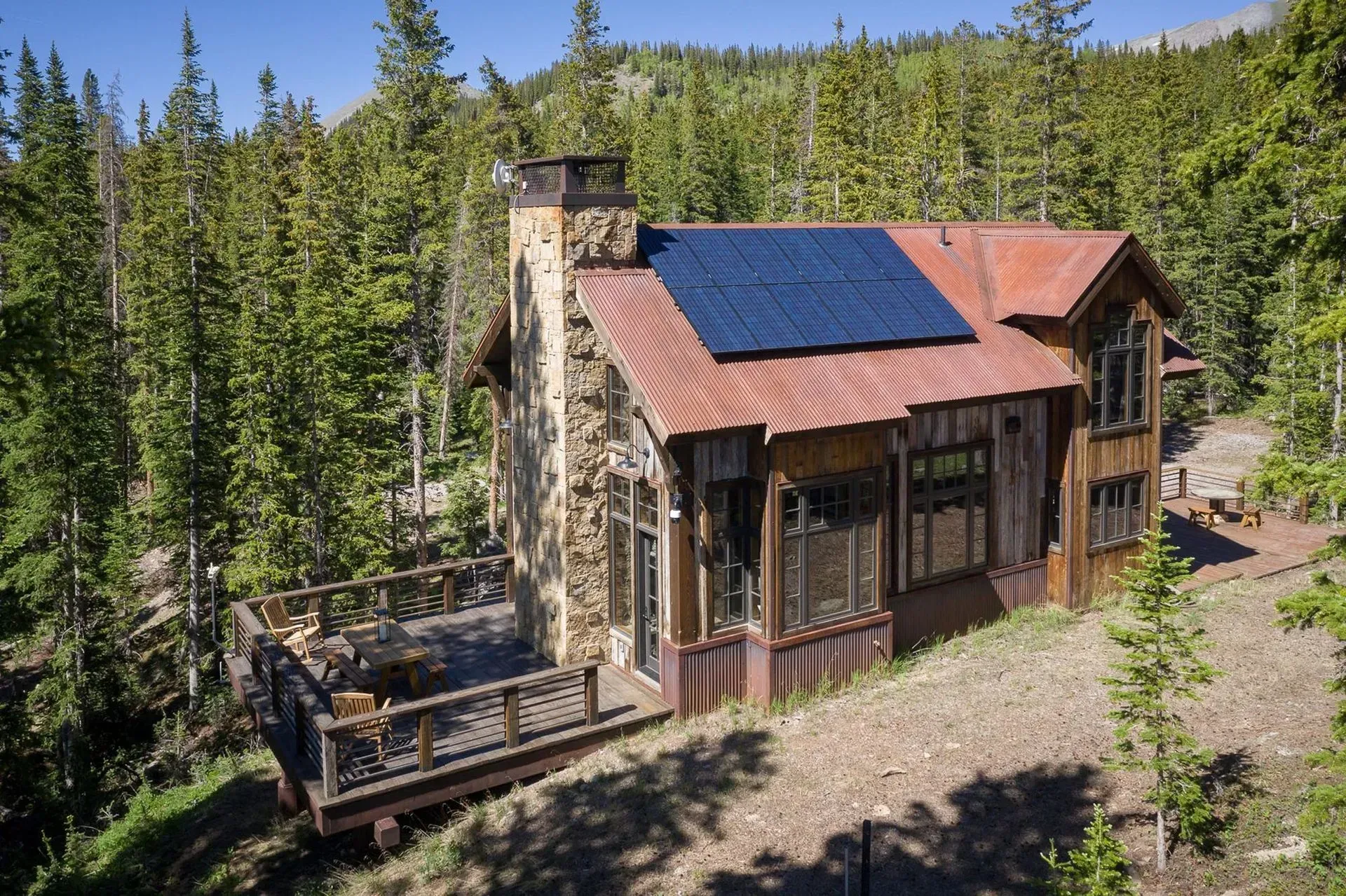Rustic cabin with solar panels, surrounded by evergreen trees, deck in foreground.