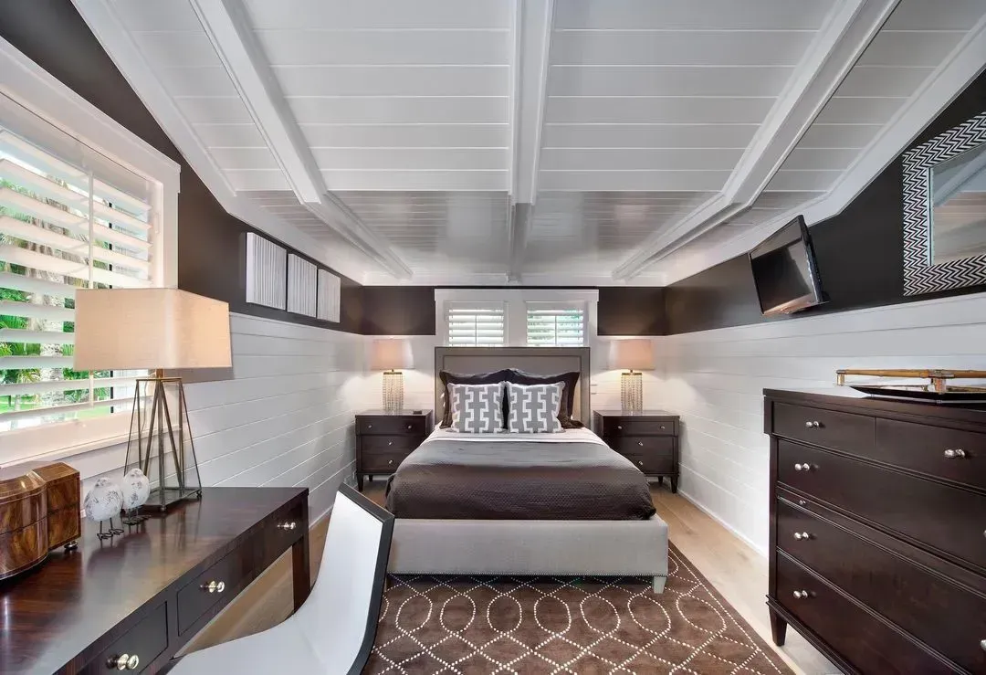 Bedroom with a bed, desk, and dresser. Neutral tones with dark brown accents and white ceiling beams.