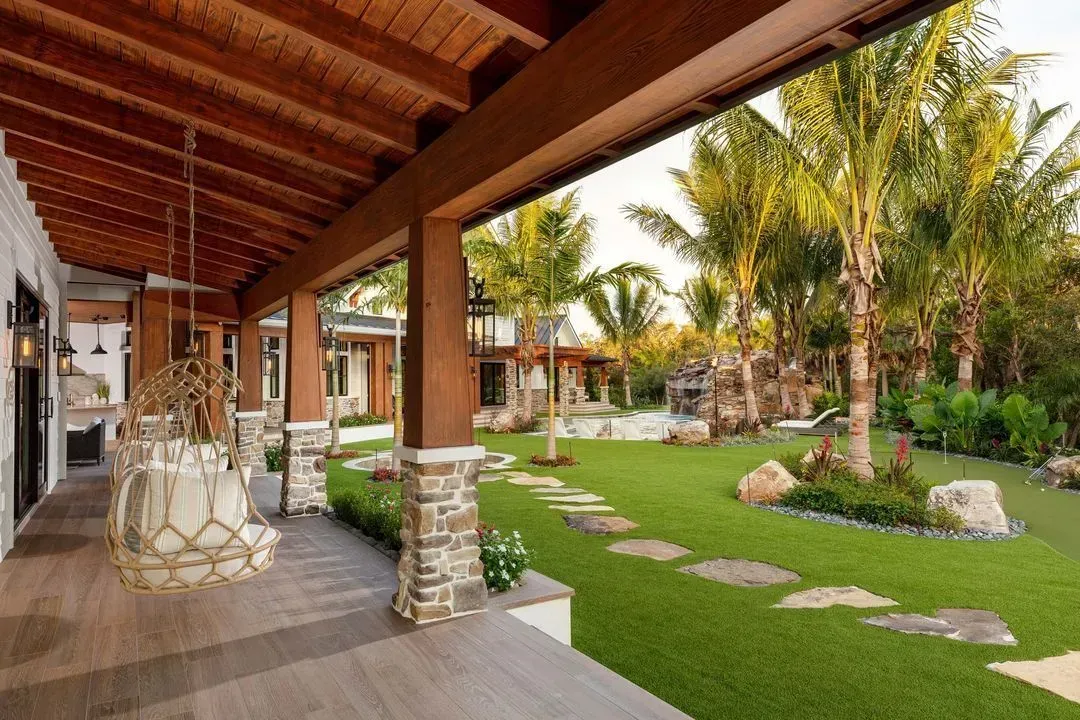 Covered porch with hanging swing overlooking a lush green yard with a pool, palm trees, and stone pathway.