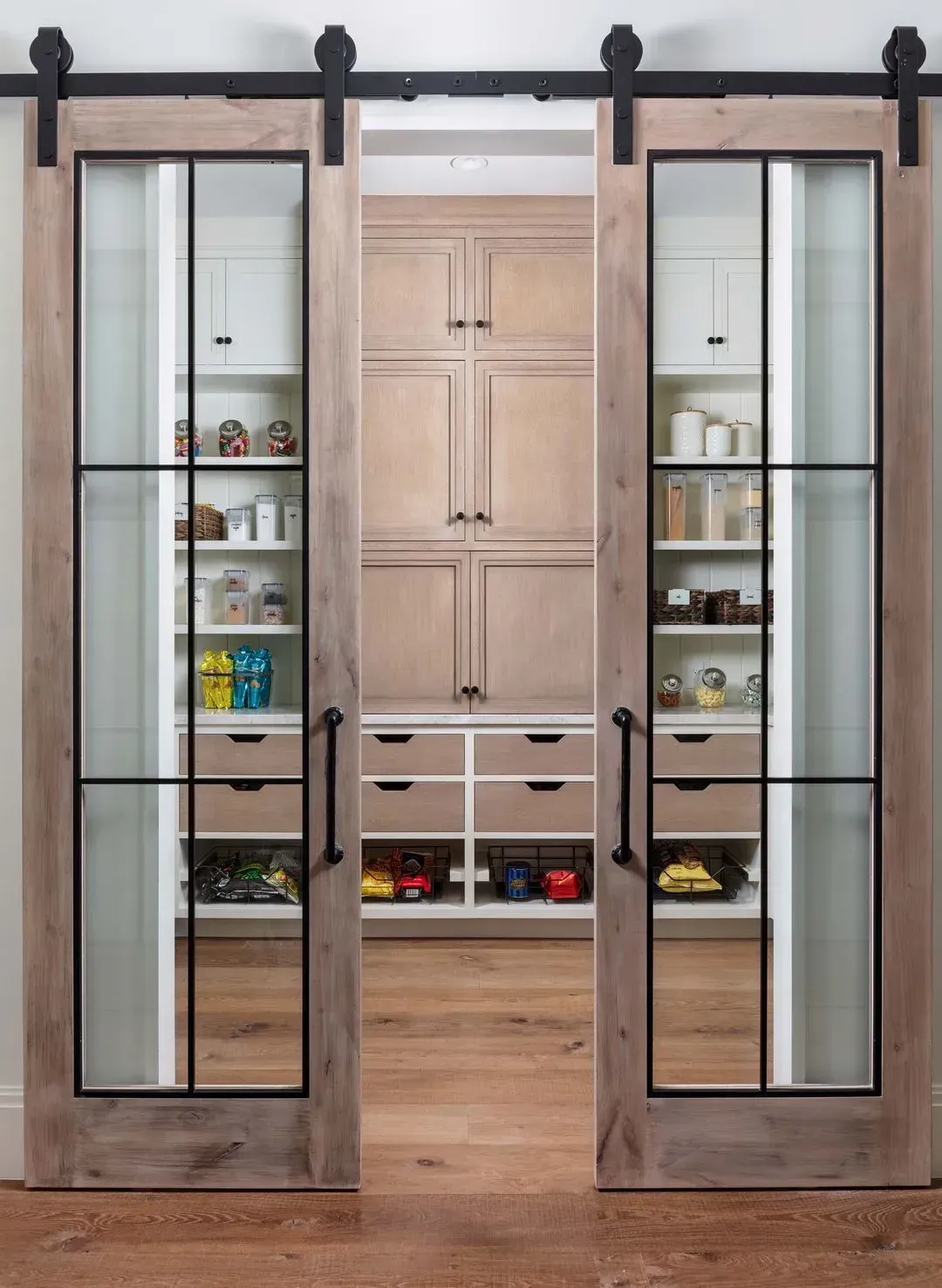 Barn doors with black frames open to a pantry with shelves and drawers filled with items.