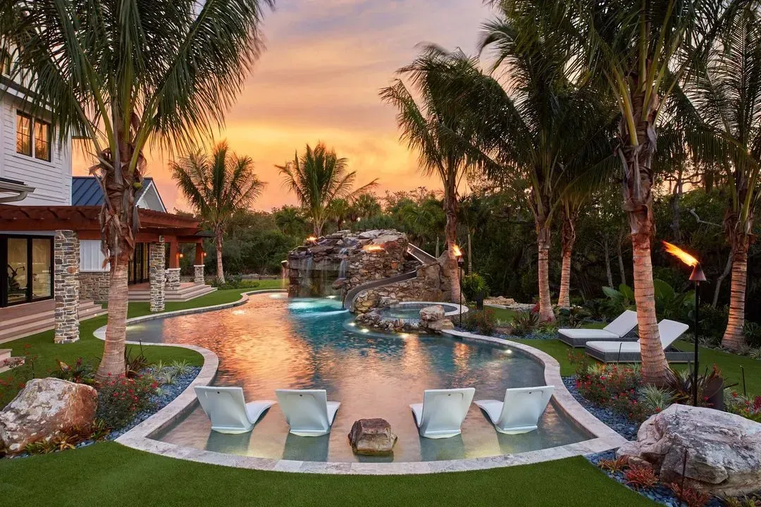Luxury backyard pool at sunset, palm trees, stone waterfall, lounge chairs.