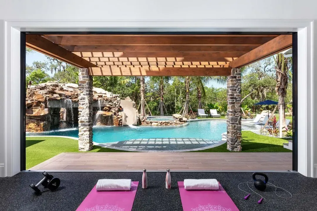 Exercise mats and weights on black floor, with a pool and waterfall view through a wooden pergola.