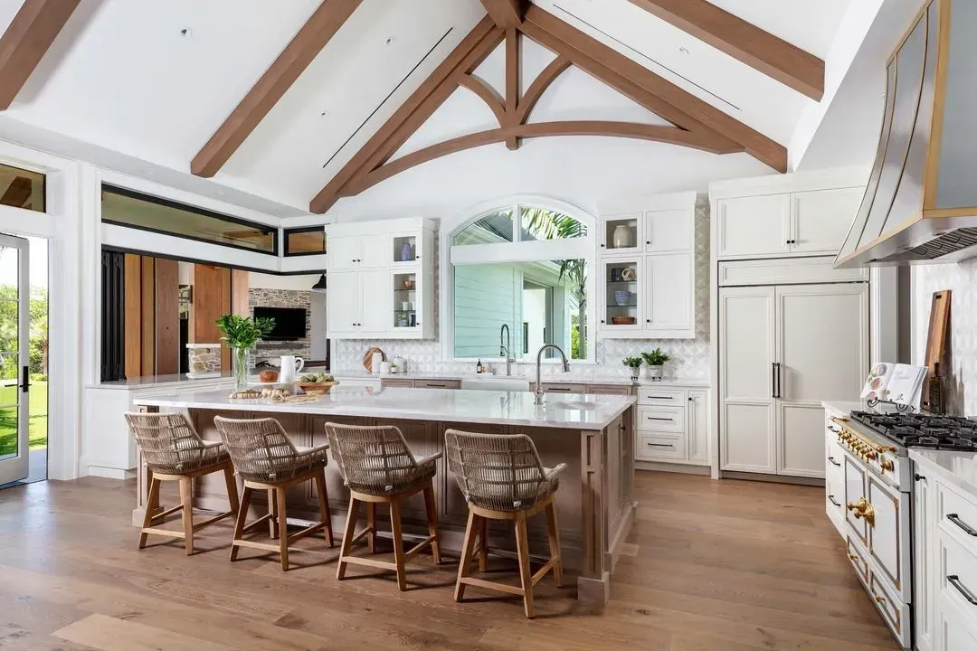 Spacious kitchen with wood beams, white cabinets, island with woven stools, and a large window.