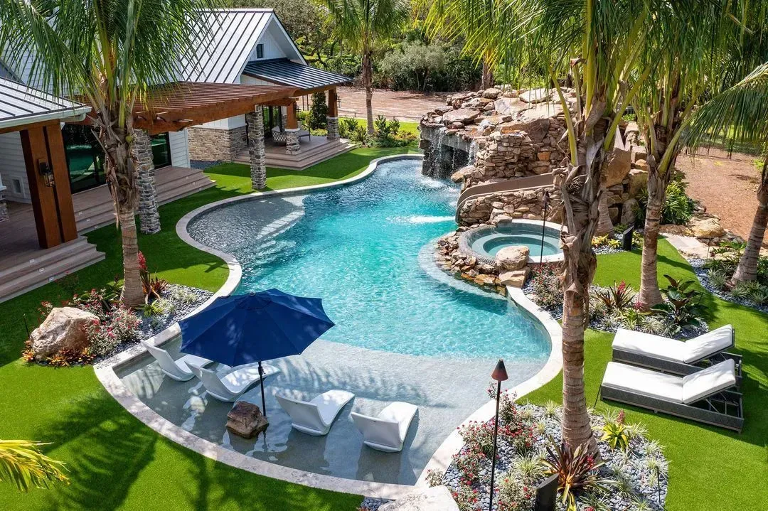 Tropical pool area with turquoise water, waterfall, jacuzzi, patio furniture, and lush landscaping.