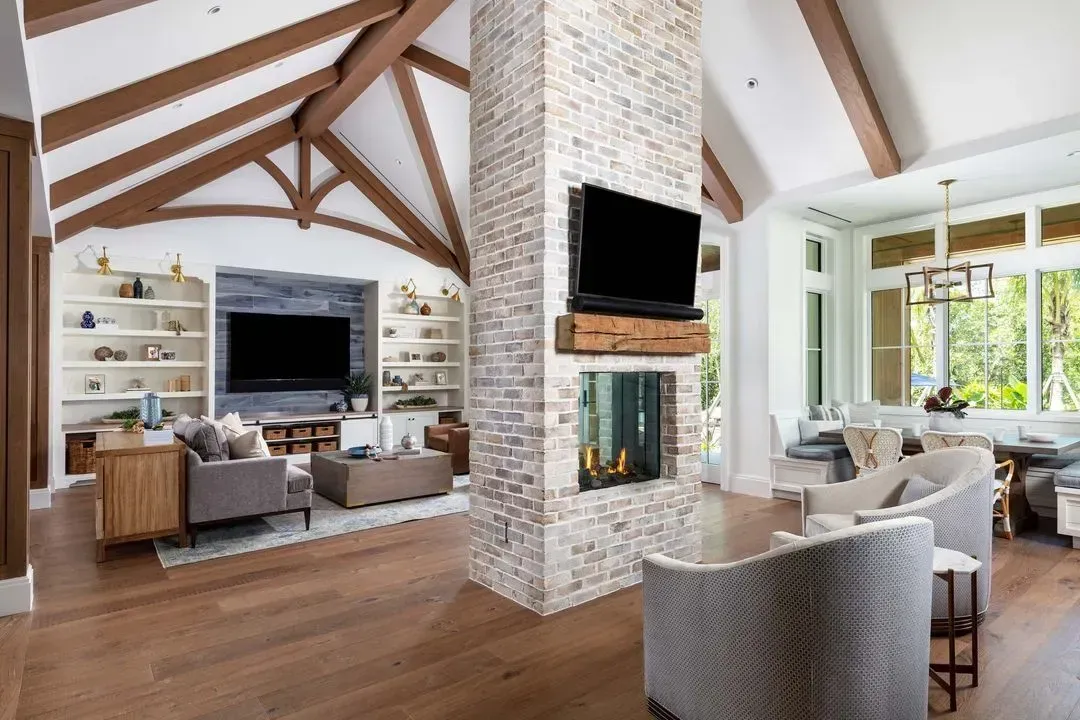 Living room with brick fireplace, TV, wooden beams, light-colored furniture, and hardwood floors.