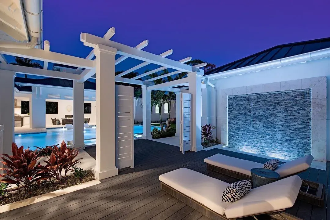 Outdoor patio with pool. White pergola, lounge chairs, stone water feature, lit at dusk.