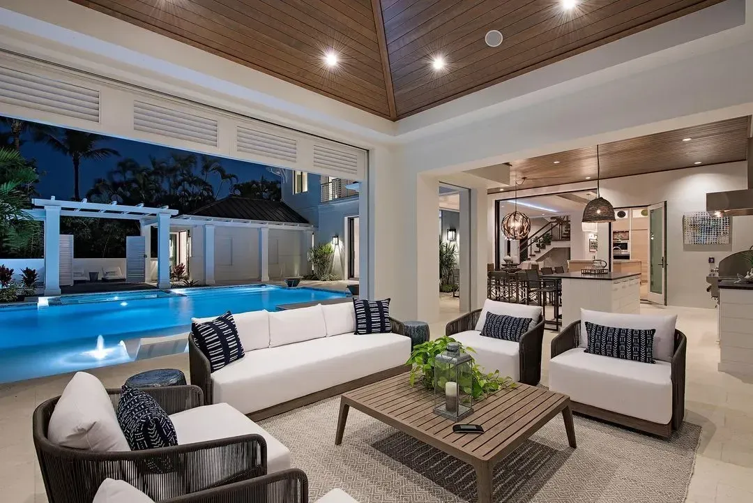 An open-air living room with pool view, white sofas, wicker chairs, and wood ceiling.