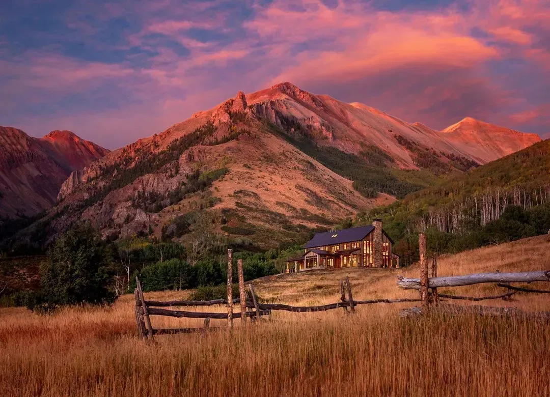 Cabin at sunset with mountains in the background, fields, and a fence, with a pink and orange sky.