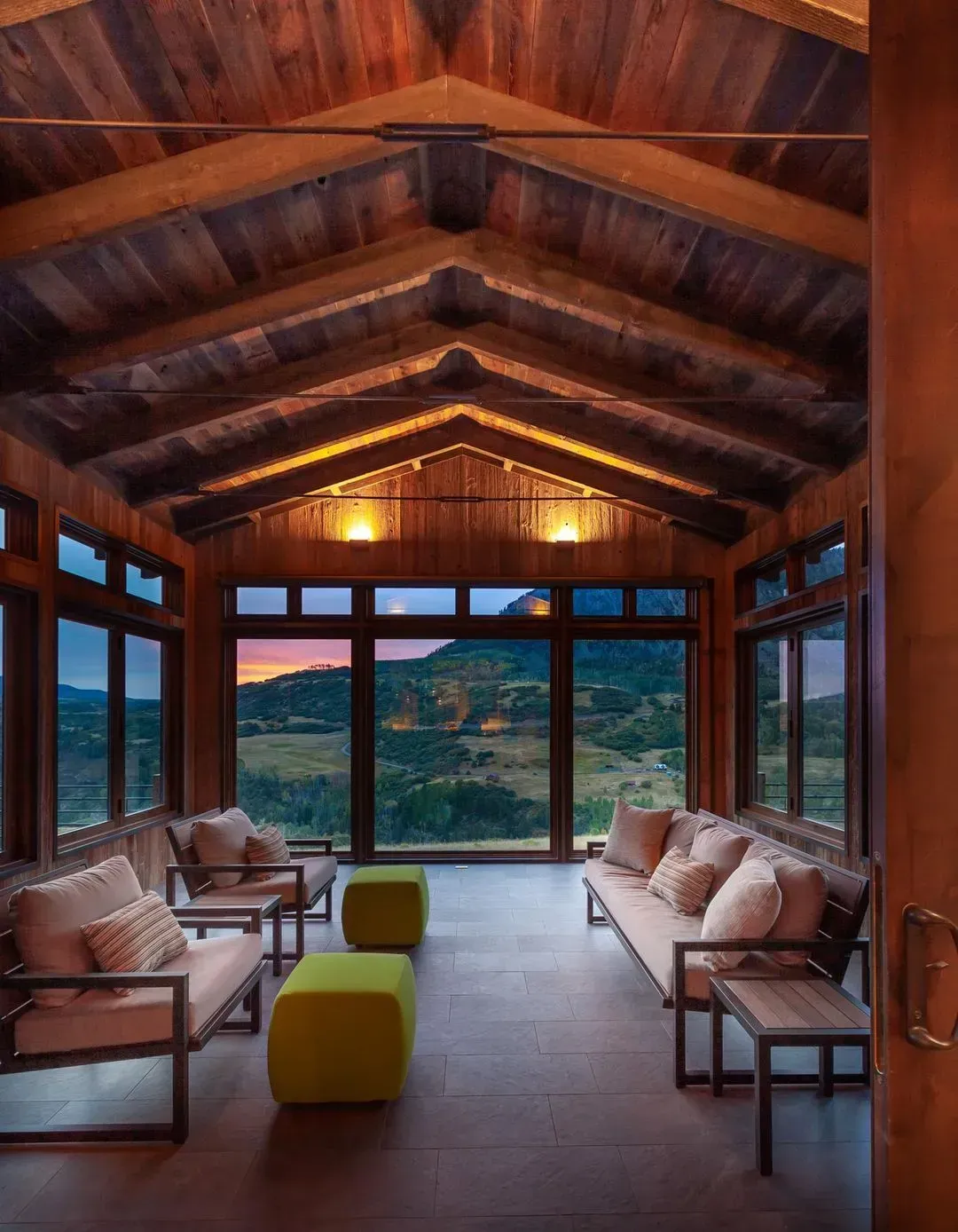 Wooden sunroom with expansive windows overlooking a landscape at dusk. Furniture includes sofas and chairs.
