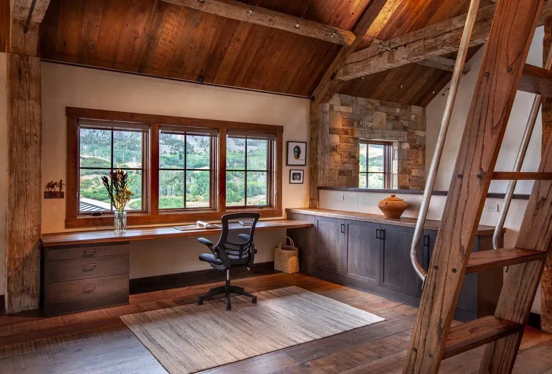 Wooden home office with desk by windows, stone wall, ladder, and rug on hardwood floor.