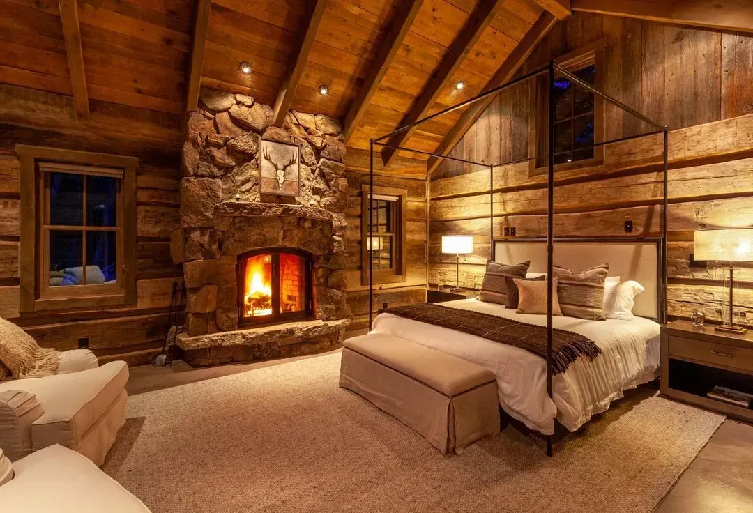 Cozy bedroom with a stone fireplace, four-poster bed, and wood-paneled walls. Fire is lit, room lit by lamps.