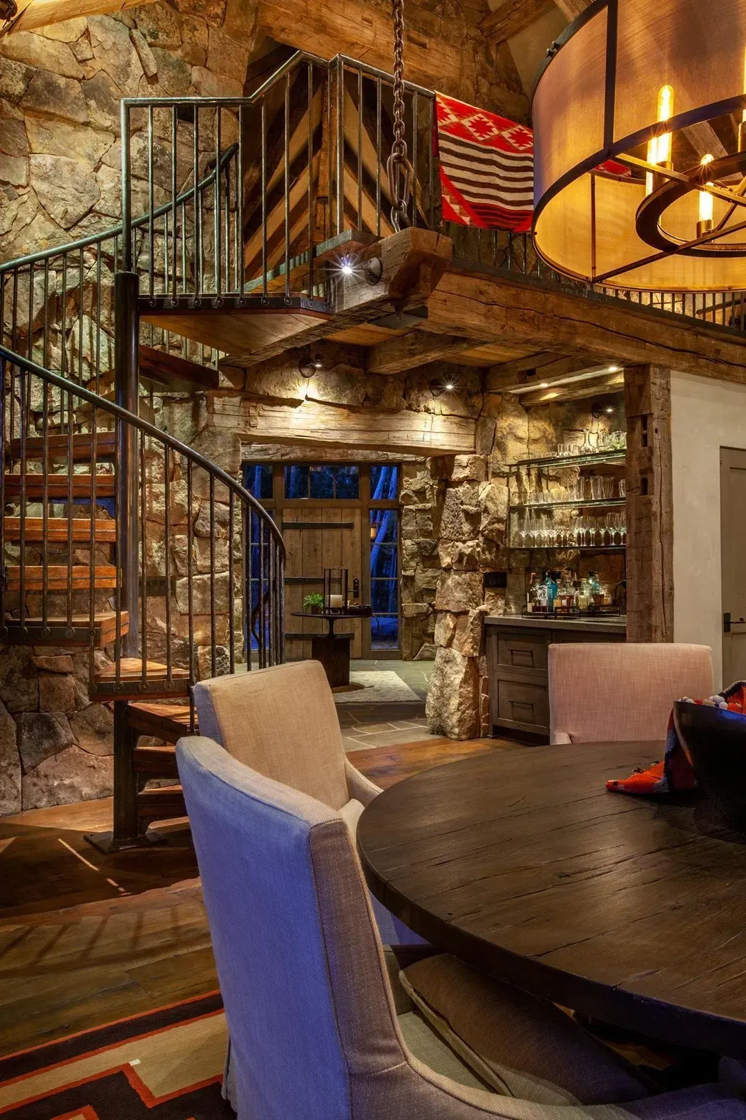 Stone-walled dining room with spiral staircase, dark wood table and chairs, chandelier.
