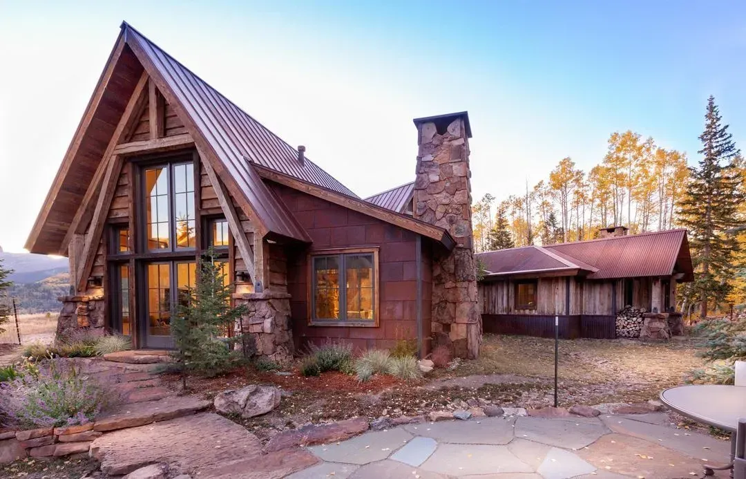 Rustic cabin with a stone chimney and A-frame roof.  Exterior features: stone, wood, and metal.