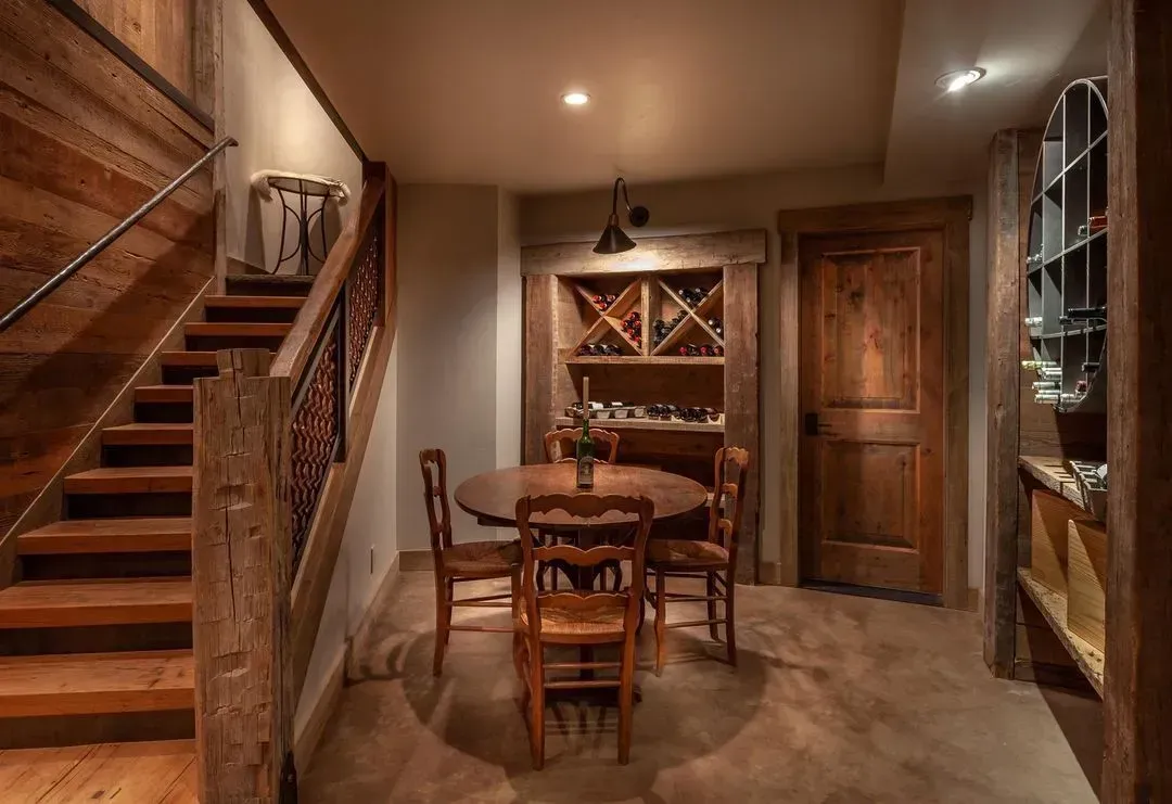 Wooden-themed dining area with a round table, wine rack, and a staircase. Warm lighting enhances the cozy space.