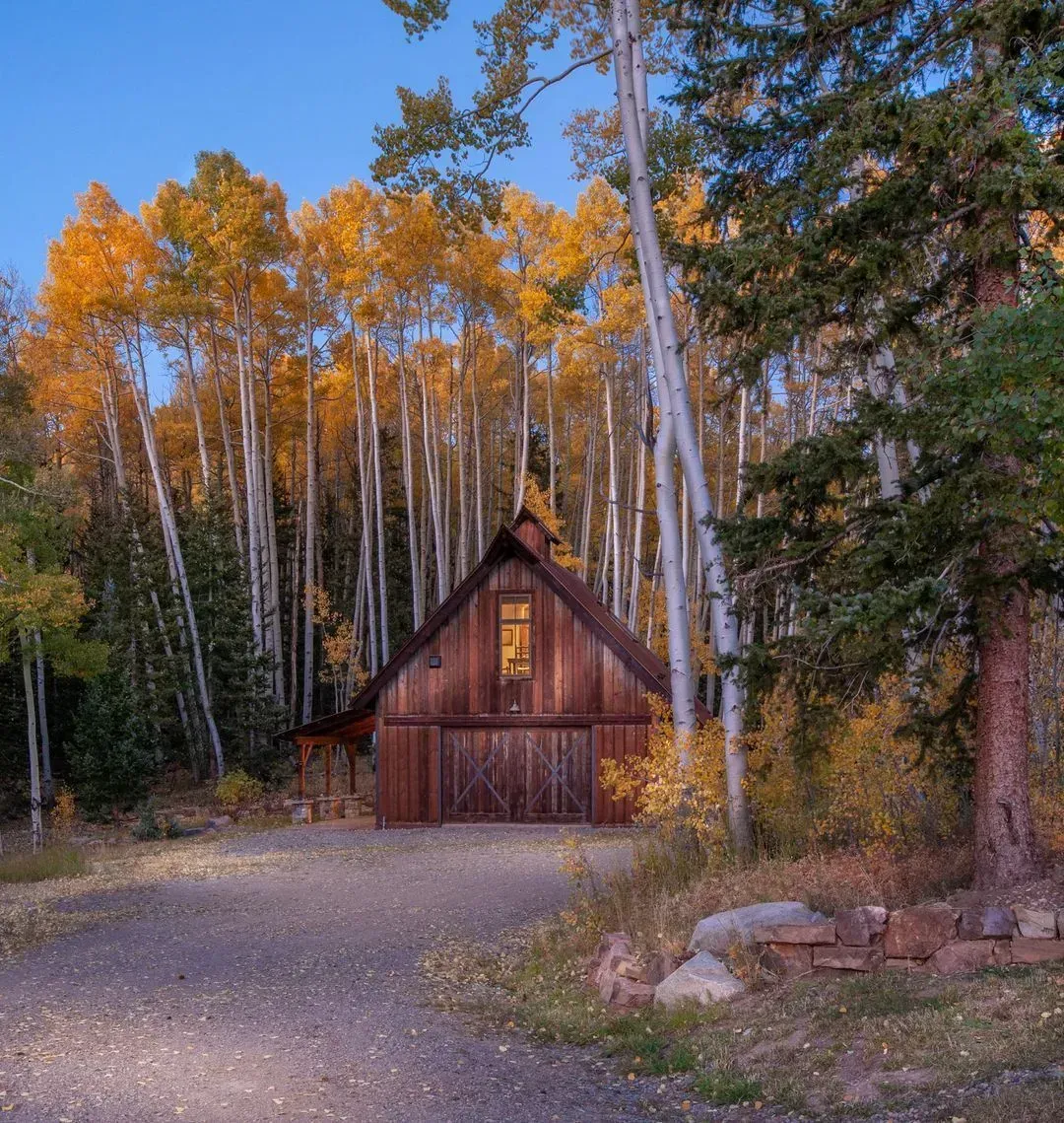 Wooden barn nestled in a forest of tall, golden-leafed trees. A dirt road leads to the barn doors.