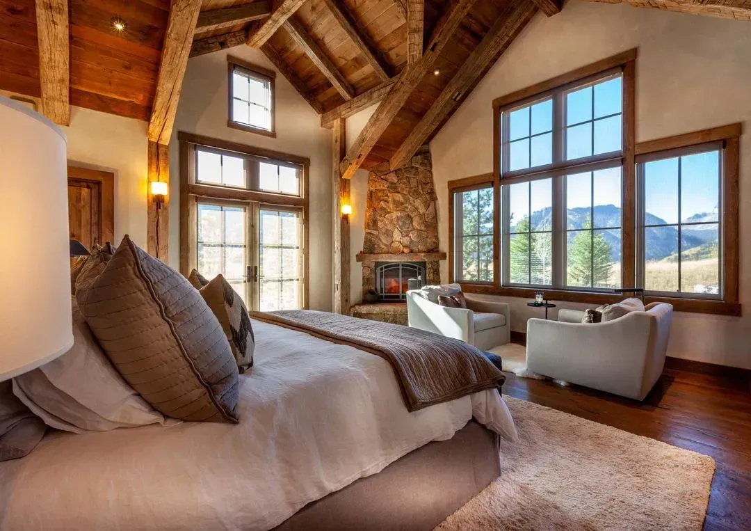 Bedroom with stone fireplace, high wood beams, large windows, and a bed.