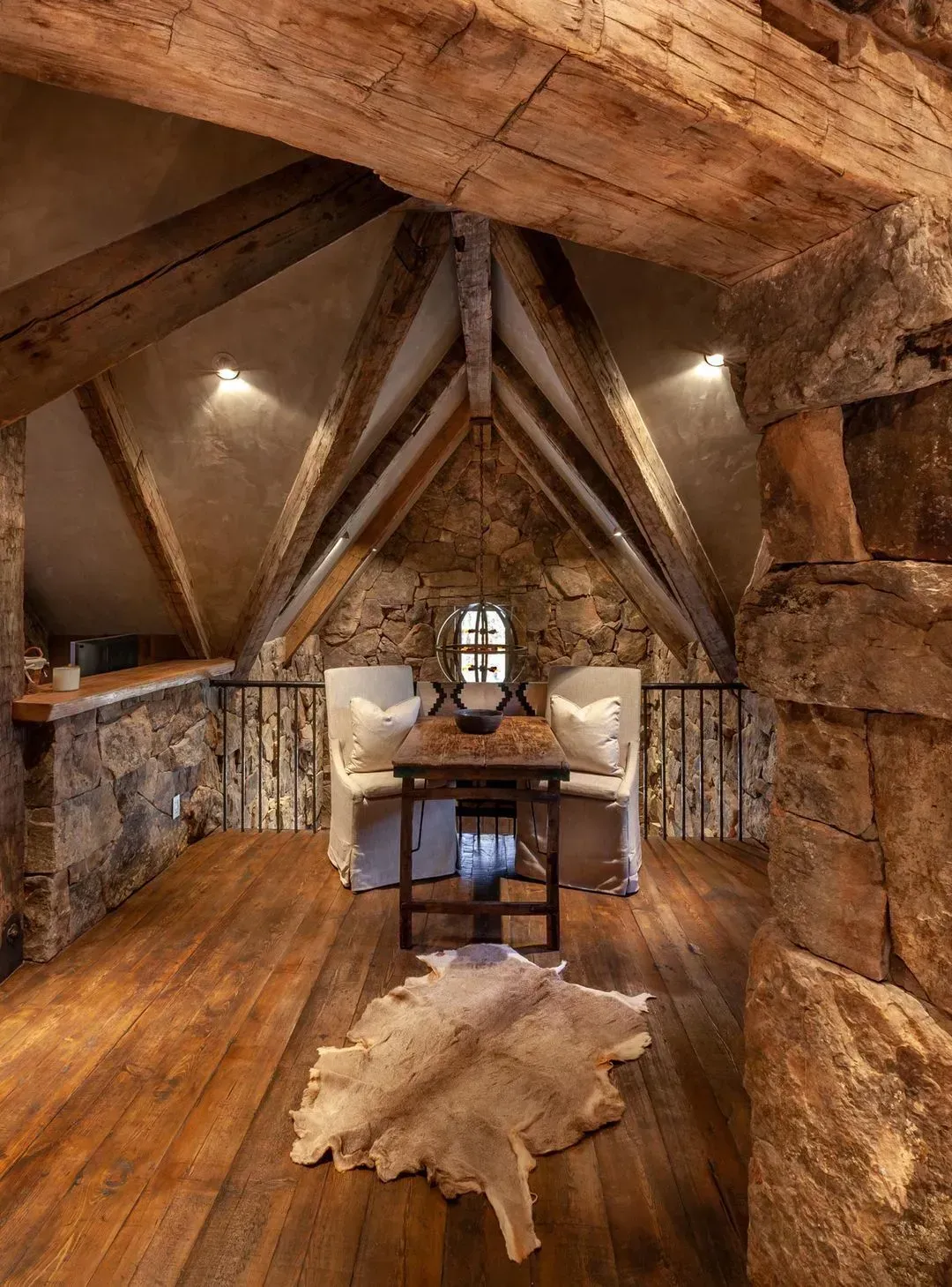 Rustic loft with stone walls, wooden beams, table, chairs, and animal hide rug.