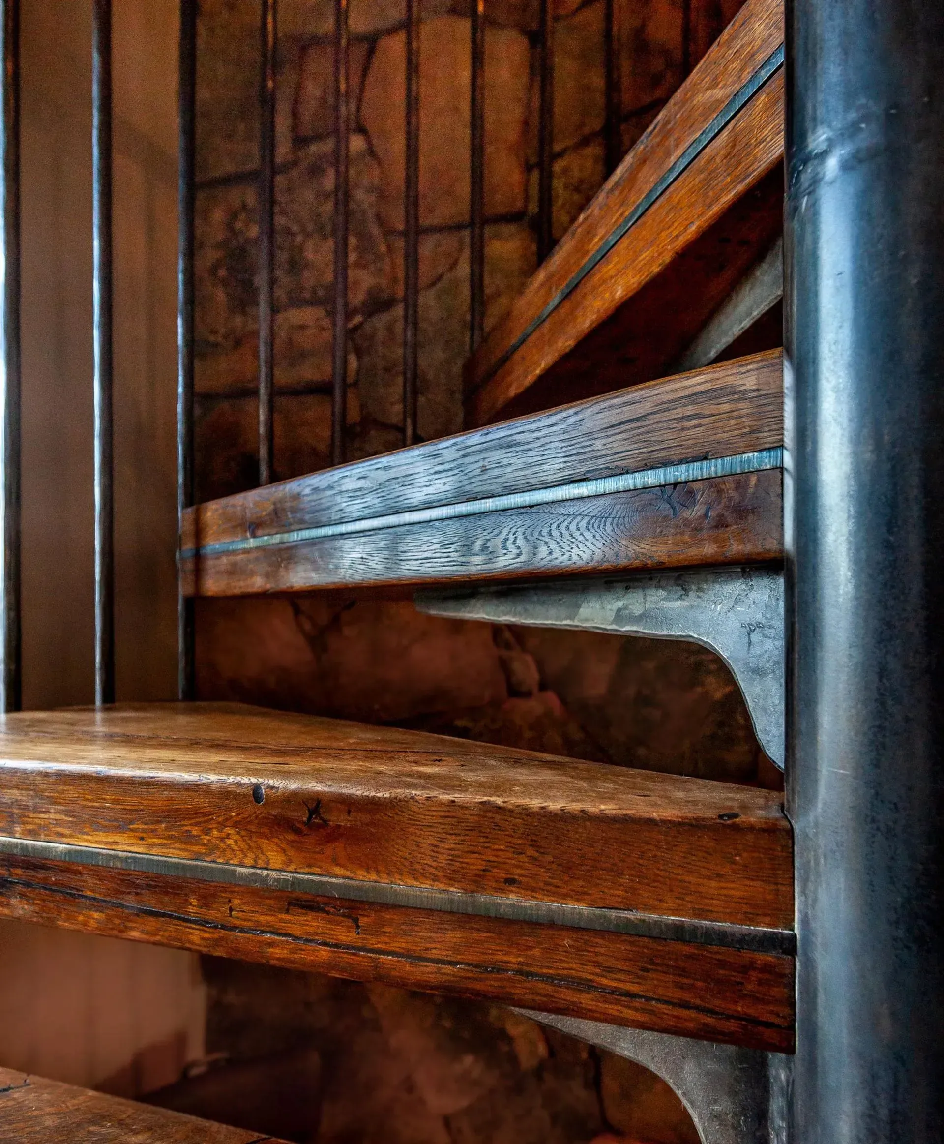 Wooden spiral staircase with metal railing against a stone wall.