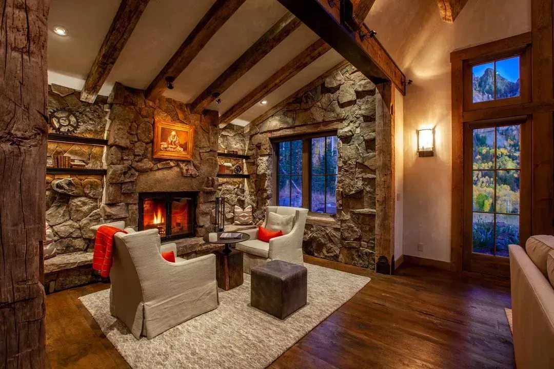 Cozy living room with stone fireplace, wood beams, two armchairs, and a rug. Evening light.